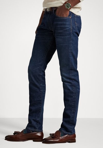 Dark blue denim jeans with a slim fit, featuring front pockets and a rolled hem, paired with brown loafers. Smooth texture, minimal design.