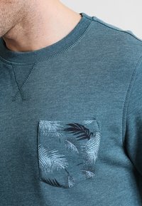 Felpa color teal con scollo tondo. Presenta un tessuto testurizzato e una tasca rettangolare con un motivo di foglie di palma in bianco e nero.