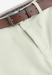Pantalons en coton beige clair avec une ceinture en cuir marron. La taille présente une doublure intérieure à motifs multicolores. Texture lisse, design classique.