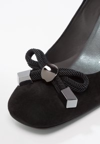 Chaussure plate en daim noir avec un nœud décoratif en cordon noir torsadé et des accents métalliques argentés rectangulaires et arrondis sur l'avant.