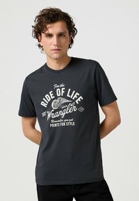Camiseta de algodón gris oscuro con un estampado gráfico blanco que incluye texto y motivos de alas. Diseño clásico de cuello redondo, mangas cortas, y corte relajado.