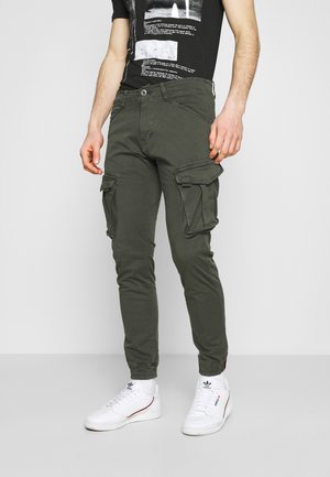 Homme portant un pantalon cargo vert foncé avec plusieurs poches, un t-shirt noir à motifs graphiques, et des baskets Adidas blanches, debout devant un fond uni.
