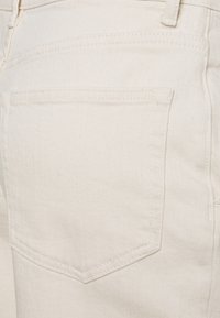 Białe dżinsy z denimu z tylną kieszenią zakończoną spiczastą klapką. Tekstura jest gładka z lekkim połyskiem, a szwy są widoczne w kontrastowym kolorze.