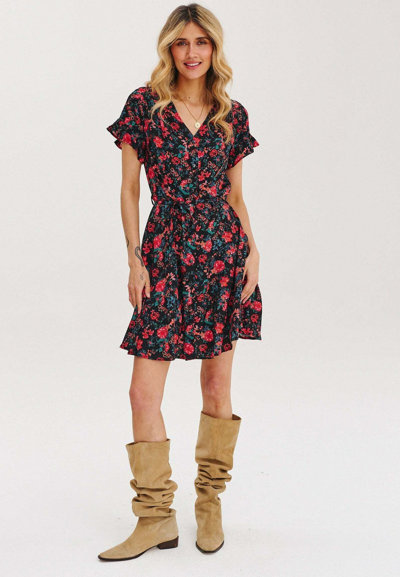 Květnatá obkládačka šatů v černé barvě s červenými a zelenými vzory, s krátkými volánkovými rukávy a zavázaným pasem. Doplněno béžovými volnými botami po kolena.