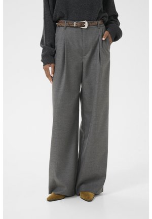 Femme portant un pantalon gris large à taille haute avec une ceinture en peau de serpent marron, un pull foncé et des chaussures pointues marron, debout avec une main dans la poche.