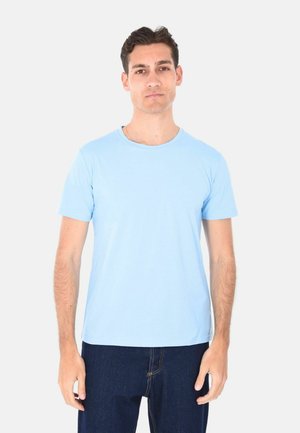 Uomo con capelli corti e scuri che indossa una t-shirt azzurro chiaro a maniche corte e jeans blu scuro, in piedi davanti a uno sfondo bianco uniforme.