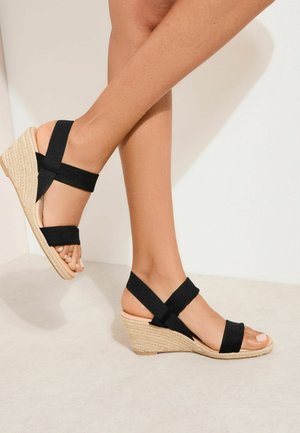 Nogi w czarnych sandałach na koturnie z materiału, z paskami wokół kostki i palców, stojące na tle jasnobeżowej ściany i podłogi.