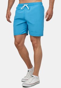 Shorts da bagno blu con cintura elastica, stringa bianca e dettaglio logo bianco di lato. Tessuto liscia e lunghezza sopra il ginocchio.