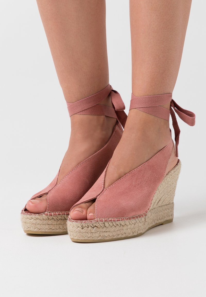 Pieds portant des espadrilles compensées en daim rose avec des rubans à nouer autour de la cheville et des orteils ouverts sur un fond clair.