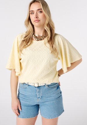 Vrouw draagt een gestructureerde lichtgele top met fluttermouwen, lichtblauwe spijkershort met witte riem en gouden ketting, poseert met hand op heup.
