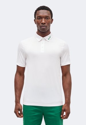 Άνδρας που φοράει λευκή κοντομάνικη πόλο μπλούζα με το «BORG» κεντημένο στο γιακά και πράσινο παντελόνι, στέκεται μπροστά σε απλό φόντο.