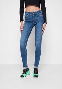 Jeans de mezclilla azul de corte slim con una cintura de altura media y detalles de desgaste ligero, combinados con sandalias de tacón bloque en negro y verde.