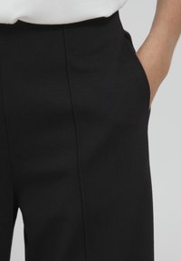 Schwarze Hose aus glattem Stoff, mit einem hohen Bund und seitlichen Taschen. Eine subtile Textur ist auf der Oberfläche sichtbar.