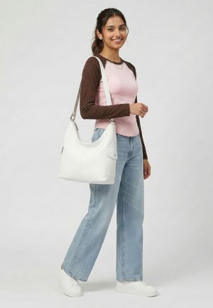 Young woman smiling, wearing a pink and brown long-sleeve shirt, light blue jeans, white sneakers, carrying a large white shoulder bag.