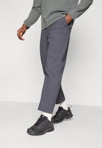 Sive hlače ravnega kroja z rahlo vezenim vzorcem blizu žepa, v kombinaciji s črnimi športnimi čevlji in belimi nogavicami.