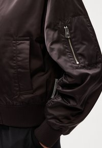 Brown bomber jacket featuring a smooth, shiny texture, ribbed cuffs, a zippered pocket on the sleeve, and a side pocket.