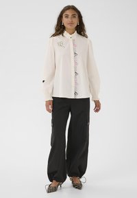 Donna che indossa una blouse bianca a maniche lunghe con ricami floreali, pantaloni larghi neri e scarpe col tacco a righe zebrate, in piedi su uno sfondo bianco.