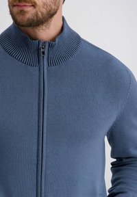 Taillierte blaue Zip-Up-Strickjacke mit einem gerippten Kragen, hergestellt aus strukturiertem Material, mit einer glatten Vorderseite und langen Ärmeln.