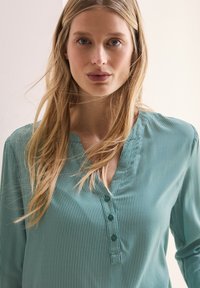 Blusa leggera color teal con righe verticali, chiusura a bottoni al collo e colletto morbido. Tessuto morbido con una leggera lucentezza.