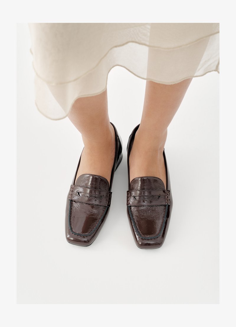 Hnědé kožené mokasíny s lesklým povrchem, hranatou špičkou, prošitými detaily a nízkým, plochým podpatkem.
