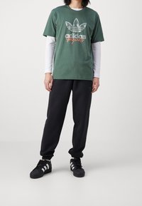 Zelené bavlněné tričko s bílými dlouhými rukávy pod ním, s vintage logem Adidas v šedé a oranžové barvě. Černé tepláky a tenisky.