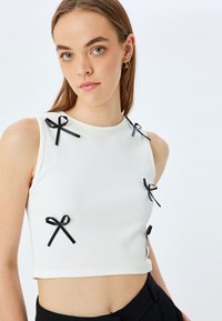 Top corto blanco acanalado con lazos de cinta negra como acentos, que presenta un escote redondo y un diseño sin mangas. Textura suave.