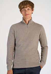Beige Zip-Hals-Pullover mit strukturiertem Strickmuster und gerippten Bündchen und Saum, getragen mit dunklen Hosen. Einfaches, sauberes Design.
