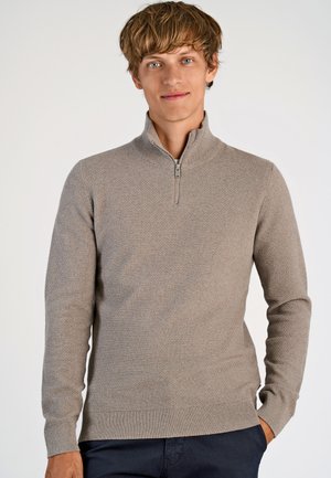 Beige ritsnecktrui met een gestructureerd breipatronen en geribbelde manchetten en zoom, gedragen met donkere broeken. Eenvoudig, strak ontwerp.