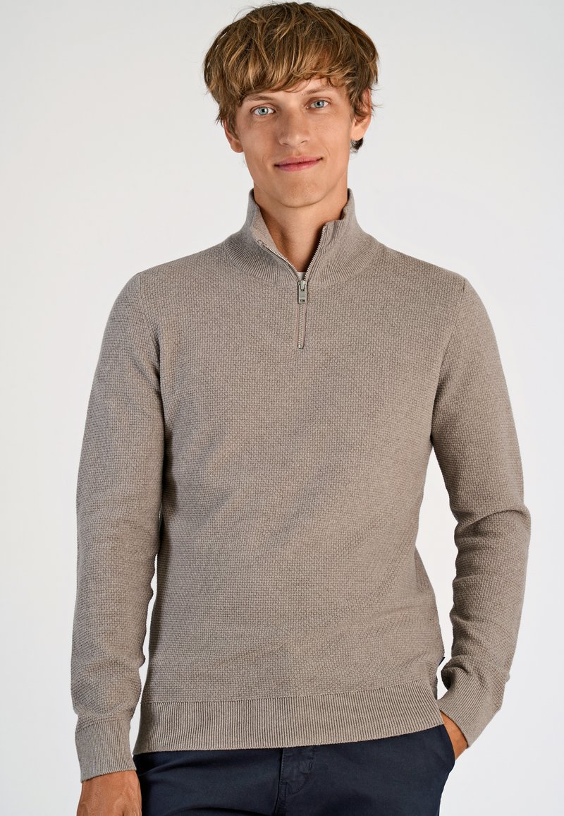 Beige Zip-Hals-Pullover mit strukturiertem Strickmuster und gerippten Bündchen und Saum, getragen mit dunklen Hosen. Einfaches, sauberes Design.