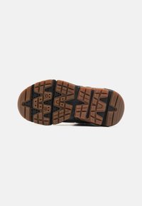 Suela de zapato deportivo negra con puntos de tracción de goma, que presenta un patrón hexagonal y acentos marrones. Texturizada para ofrecer agarre y durabilidad.