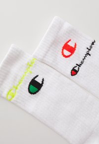 Calcetines de algodón blancos de caña media con tejido texturizado. El calcetín izquierdo presenta un logo bordado en amarillo neón y verde; el calcetín derecho tiene un logo rojo.