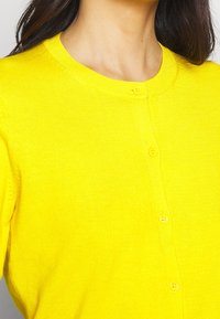 Gelbes Strickcardigan aus Knitstoff. Verfügt über einen runden Ausschnitt und sechs Vorderknöpfe. Glatte Textur mit taillierter Form.