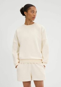 Crèmekleurige sweatshirt met een losse pasvorm, voorzien van geribbelde manchetten en zoom. Gecombineerd met bijpassende crèmekleurige shorts, wat zorgt voor een samenhangende look.