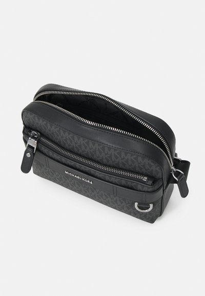 Sac en toile noire avec double zip, détail de logo embossé et accents en cuir. Présente une forme structurée et une texture contrastée.