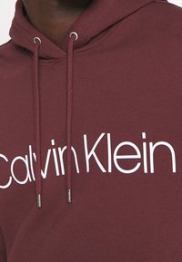 Bordový mikina s kapucí a bílým logem "Calvin Klein", tkaničky se stříbrnými konci a žebrovanou texturou na manžetách a lemu.