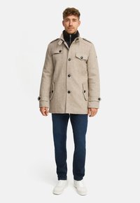 Beiger Mantel aus Wollmischung mit doppelreihigem Design, vier vorderen Taschen und Knopfdetails. Kombiniert mit dunklen Jeans und weißen Sneakers.