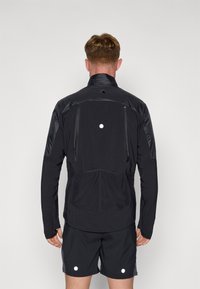 Schwarze Sportjacke mit hohem Kragen, Reißverschluss-Taschen und reflektierenden Akzenten. Der Rücken hat ein elegantes Design mit einem gerafften Bund.