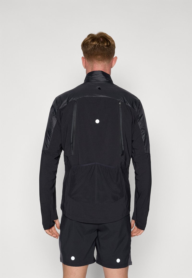 Chaqueta deportiva negra con un cuello alto, bolsillos con cremallera y detalles reflectantes. La parte posterior tiene un diseño elegante con una cintura fruncida.