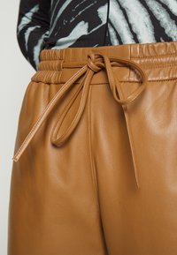 Brune læderbukser med en bundet snøre i taljen, båret med en sort og hvid mønstret bluse.