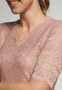 Spitzenbluse in sanftem Rosa, mit einem gewellten V-Ausschnitt und kurzen Ärmeln, die mit aufwendigen Blumenmustern und einer strukturierten Oberfläche verziert sind.