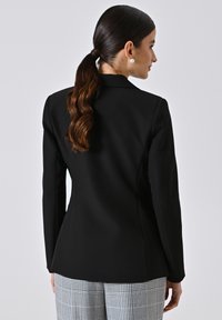 Zwarte op maat gemaakte blazer met een gladde textuur en gestructureerde pasvorm, met een enkele split aan de achterkant, gecombineerd met broek in ruitpatroon.