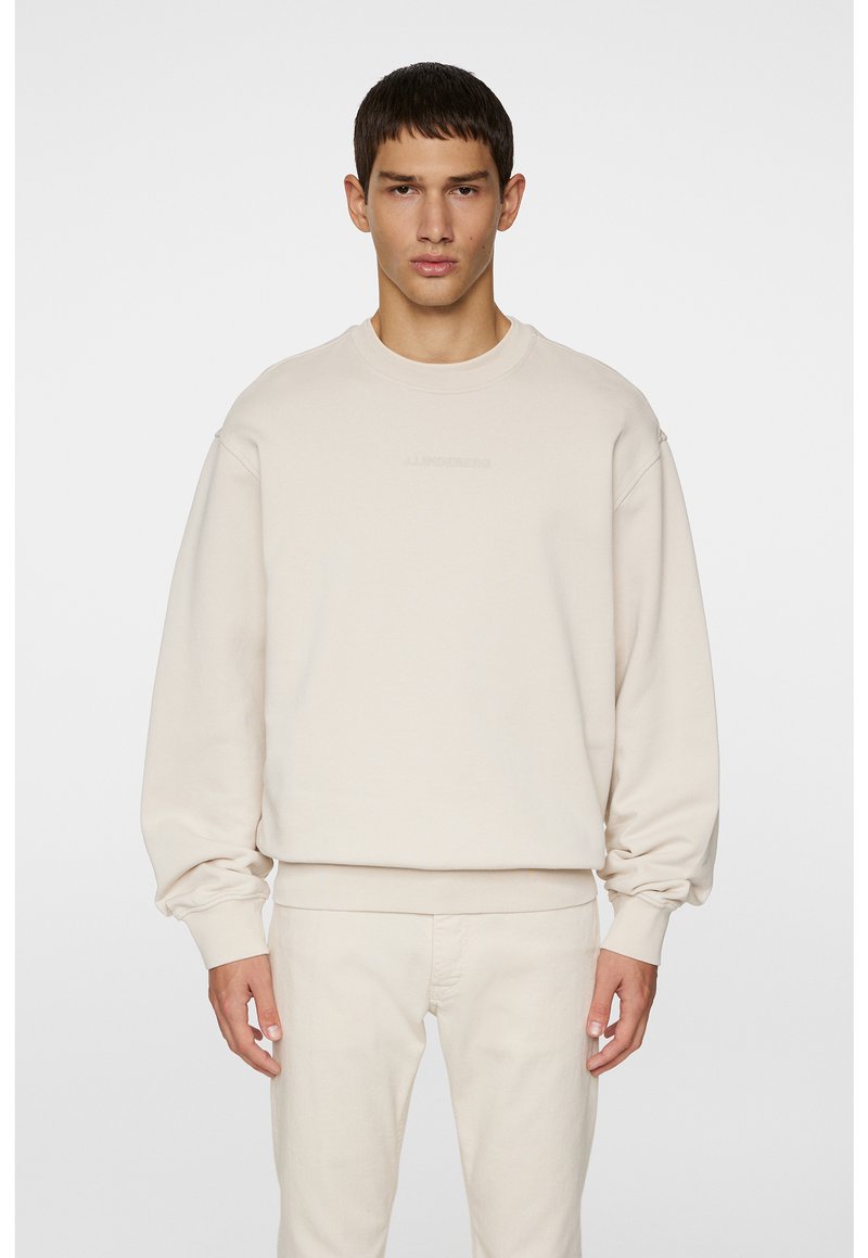 Beige sweatshirt med rund halsudskæring, sænkede skuldre og ribbede manchetter. Har diskret logo-detaljering på brystet. Blød stoftekstur.