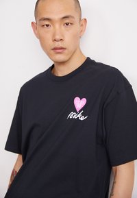 Juodos medvilnės T-palaidinė su apvaliu kaklu, kuriame yra rožinės širdies grafika ir "Nike" tekstas baltu spasnyba, turi trumpas rankoves ir laisvą prigludimą.