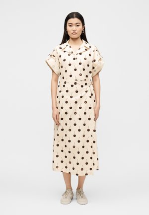 Žena stojaca, oblečená v krémovom midi šatách s vyhrnutými krátkymi rukávmi a vzorom čerešní, v kombinácii so svetlo béžovými teniskami.