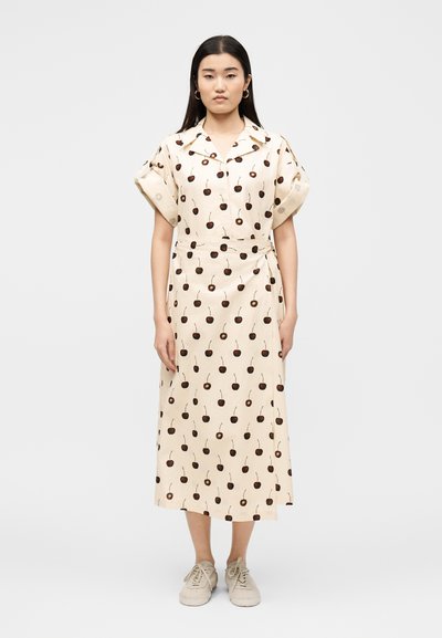 Mujer de pie, vistiendo un vestido midi color crema con mangas cortas remangadas y un estampado de cerezas, combinado con zapatillas deportivas beige claro.