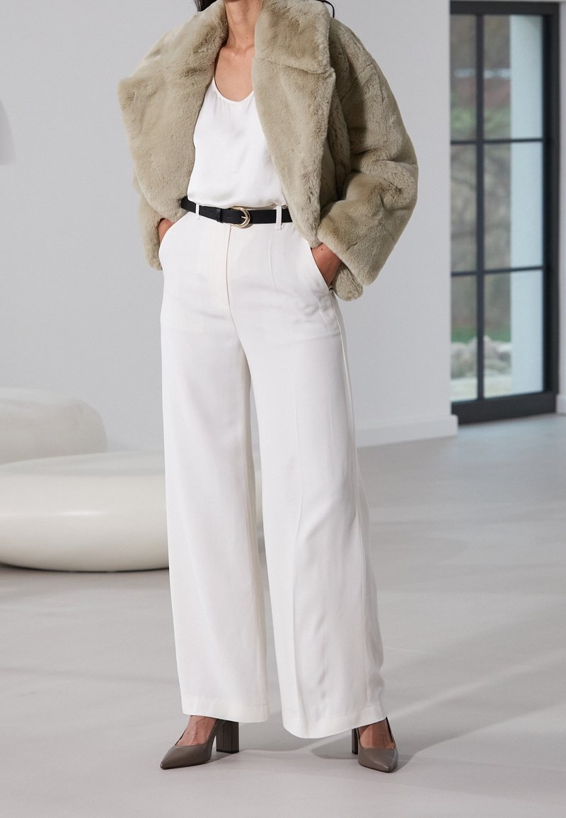 Femme portant une veste en fausse fourrure beige, un pantalon blanc à jambes larges, un haut blanc, une ceinture noire et des chaussures à talons gris, debout à l'intérieur.