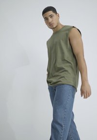 Canotta verde oliva senza maniche con una vestibilità comoda, abbinata a jeans dritti blu chiaro, su uno sfondo bianco semplice.