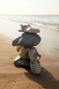 Valkoiset muoviset sandaalit, joissa on hihnallinen muotoilu, nilkkaremmi ja solki, asetettuna sileiden kivien päälle rannan läheisyydessä.