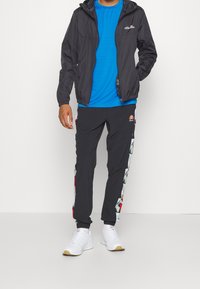 Veste imperméable noire avec capuche, t-shirt bleu à évacuation d'humidité, pantalon noir avec des motifs latéraux colorés, et chaussures de sport blanches.