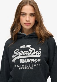 Sudadera negra con el texto "Vintage SuperDry" en blanco, hecha de un material de algodón suave, ajuste holgado y un acogedor bolsillo tipo canguro.